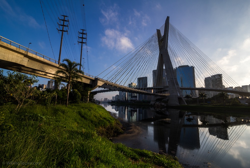 Essa imagem está aqui para você entender melhor como é a margem do rio e o que esperar quando estiver lá para fotografar a Estaiada. Na imagem não é possível ver, mas a ciclovia está à esquerda. Em alguns lugares, a região bem próxima ao rio é mais plana e menos perigosa de você cair. Se você se aproximar do rio, tome extremo cuidado para não cair nele! Nessa imagem em baixa resolução talvez você não consiga perceber, mas tem dois amigos ali perto da margem e da ponte, vamos ver como fica a vista de lá na próxima foto! | ISO 100; 16mm equiv.; f/11; composição com tempos de exposição diferentes